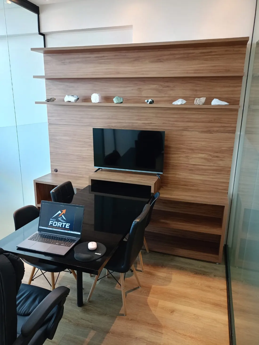 Sala de reunião com TV, mesa preta e notebook com o logotipo Rio Forte